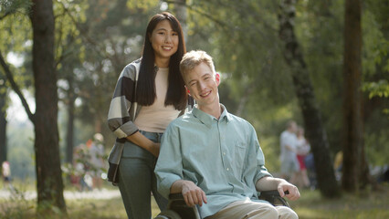 A cute asian lady with long dark hair and her disabled young man in a blue shirt are standing in the public park, looking at the camera and smiling