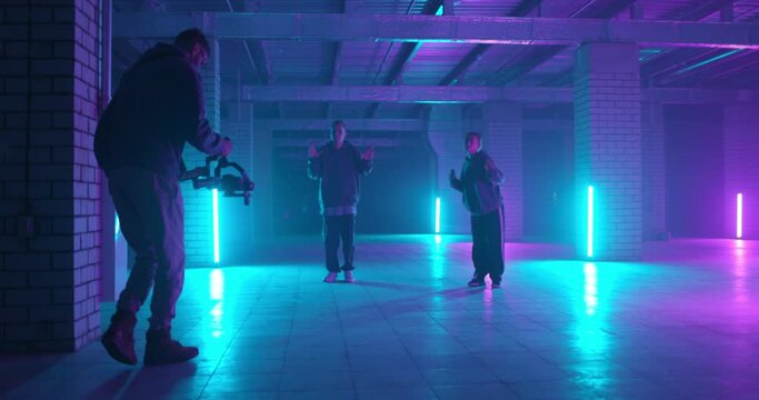 Backstage of video shooting. Cameraman filming hip-hop dancers in neon light. Operator moves with image stabilizer in hands at dark studio. Shooting a clip