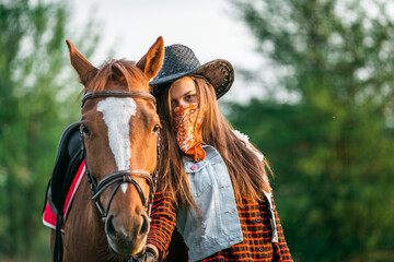 A cowgirl stands near her horse in a field at sunset.