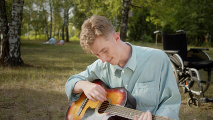 A young disabled blonde man is sitting on the plaid in the public park and playing guitar professionally 