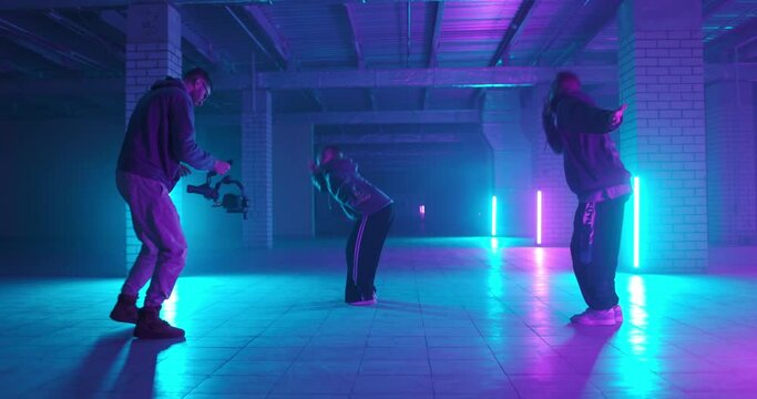 Backstage of video shooting. Cameraman filming hip-hop dancers in neon light. Operator moves with image stabilizer in hands at dark studio. Shooting a clip