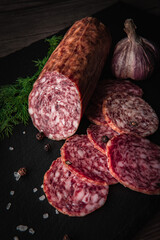 Sliced salami sausage on a board. Meat product and herbs on a dark back. A quick meal or snack. Dried sausage with black peppercorns.