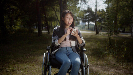 Fototapeta na wymiar A pretty girl is sitting in a wheelchair in a public park and looking into the screen of her mobile phone, searching through the Internet and smiling