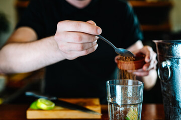 hands of a young waiter, adding brown sugar in a large crystal glass to prepare a Mojito. horizontal.