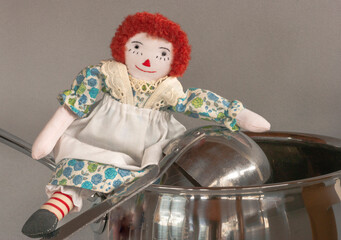Raggedy Ann with apron leans on soup ladle and kettle as if getting ready to cook. Size comparison is shown by miniature doll with large scale pot in closeup.