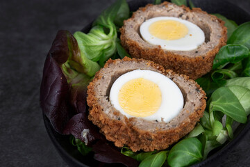 Scotch egg halved on lettuce leaves in a black dish.  On a stone background