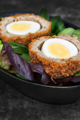 Scotch egg halved on lettuce leaves in a black dish.  On a stone background