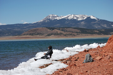 An angler reclines in a snowbank wait for a trout with mountains in the background.  
