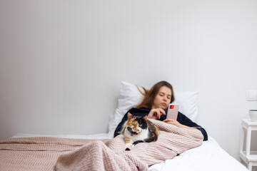 Pets, morning, comfort, vacation and people concept - young woman looking at phone and she has a tricolor cat sitting on her feet.