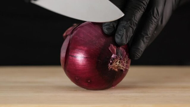 One red onion lies on cutting board. Large kitchen knife cuts Italian red Tropea onion into two pieces. Human hands in black gloves cuts onion in half with on black background. Close-up slow motion