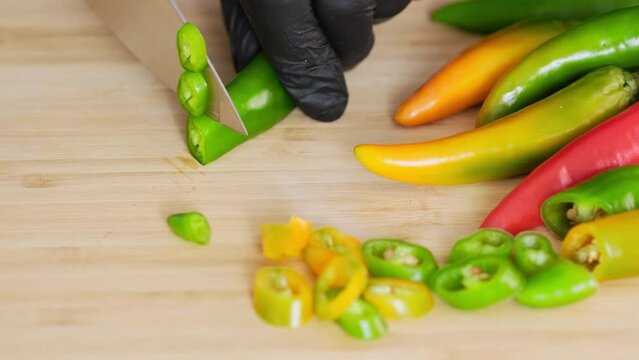 Hot chili peppers. Chef slicing jalapeño peppers on chopping board in kitchen. Cutting or chopping whole red yellow green fresh pepper with sharp knife into pile of slices