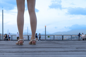 long legs of young lady with sandals standing on wooden floor of a pier in Hong Kong. evening cloudy sky in background