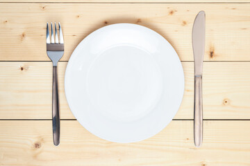 Top view of an empty white plate, fork and table knife on a wooden background