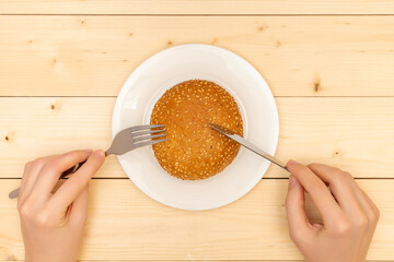 Unhealthy food. Female hands with a fork and a table knife carve a sesame bun, top view