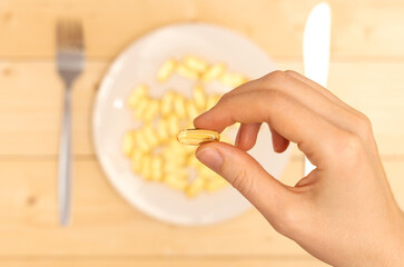 One capsule of vitamin omega 3 in a woman's hand, a handful of capsules on a white plate in the background. Selective focus.
