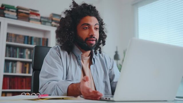 Young attractive Arabian man with headset on head participates in online rally looks at laptop internet interview with corporation to get good offer sits at table in home office