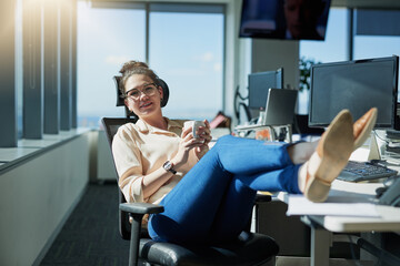 Work is so laid back today. Shot of a cheerful young businesswoman drinking coffee while sitting with her feet up on her desk inside of the office.