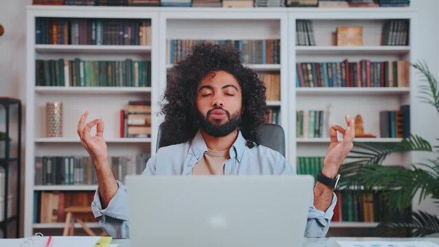 Young pacified Arabian man office worker does breathing exercises yoga in lotas position and closes eyes to relax or restore strength meditates after hard day work sits at table in front of laptop