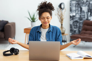 Young African American woman sitting at the office desk with a laptop meditating eyes closed...