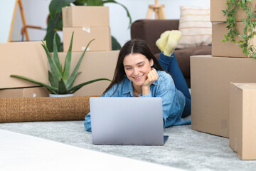 Moving to a new house, rental housing. Happy young caucasian woman using laptop computer to search and order a transportation service and movers to move to a new home