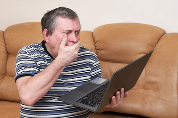 A man with a surprised face holds a laptop in his hands while sitting on couch - 496576257