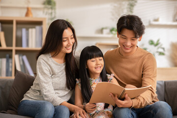 Happy Asian family sitting on the sofa together looking at daughter's note book. Parents are smiling, reading daughter book and proud of education progress. Watching book together, family relationship
