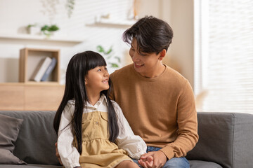 Smiling Asian father embracing daughter sitting on couch, posing for family photo indoors. Happy young dad with little child girl, resting on sofa in the living room.