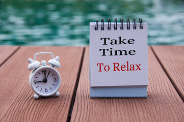 Alarm clock on wooden floor with text on notepad - Take time to relax. With blurred swimming pool background. The clock set at 9 o'clock.