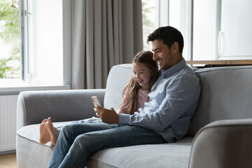 Obraz premium Happy excited young dad and cute daughter using online learning app on smartphone, sitting on couch at home, speaking on video call, laughing, smiling, enjoying communication