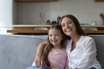 Happy loving young mom hugging cheerful little daughter girl, feeling joy, tenderness, bonding, affection, enjoying motherhood, leisure, warm family relationship, looking at camera away, smiling