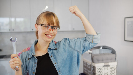 Independent caucasian woman in a jean shirt holding a wrench tool, looking at camera with one hand up, showing off her muscles. High quality photo