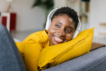 Smiling multiracial woman lying on sofa and listening music in headphones. Girl enjoying audio...