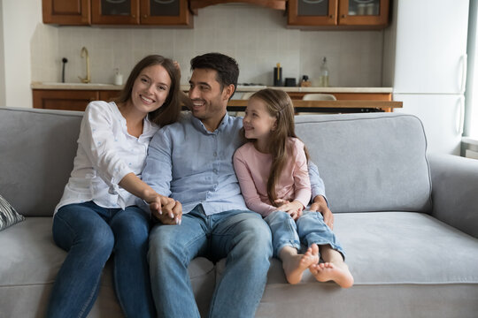 Happy loving young parents and preschool little kid home portrait. Cheerful dad and cute daughter girl looking at mom, hugging, Mother posing on couch near father and child. Family, parenthood concept