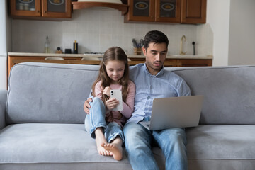 Focused gadget addicted young dad and little gen Z daughter girl engaged in laptop computer, smartphone using. Father and kid sitting on sofa at home, browsing internet, social media, playing games