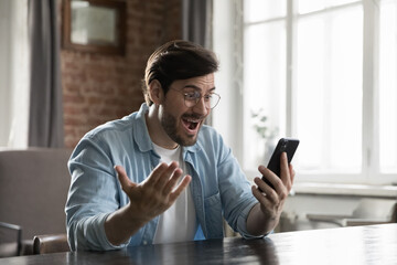 Excited smartphone user man staring at screen in surprise, reading text message getting good news,...