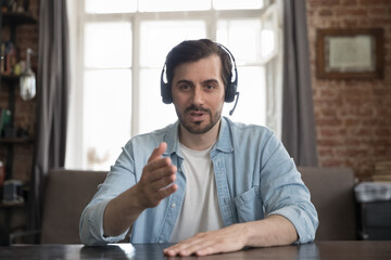 Confident millennial freelance employee man in headphones with mic head shot portrait. Positive engaged blogger talking on video conference call, speaking at mic, looking at camera