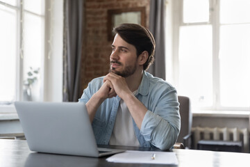 Thoughtful dreamy business professional man working at laptop in home office, looking at window,...