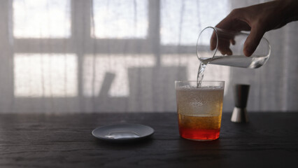 pour tonic water over ice ball with orange syrup in tumbler glass on wood table