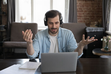Positive busy millennial freelance employee man in headset speaking at webcam on laptop computer,...