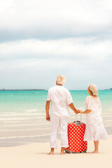 Caucasian senior travellers in white clothes outdoors on a beach with suitcase