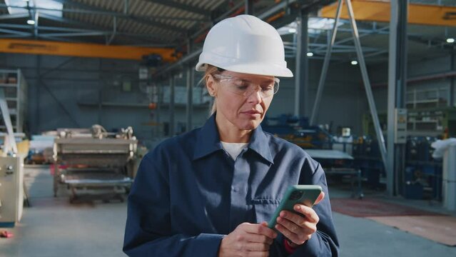 Portrait serious woman with helmet, uniform and goggles uses a mobile phone in factory. In the background details of a metalworking project
