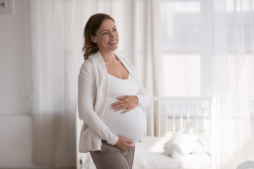 Happy cheerful expecting mom touching big baby bump, looking away, dreaming of motherhood. Young pregnant woman thinking of childbirth, smiling at good thoughts. Pregnancy, maternity concept