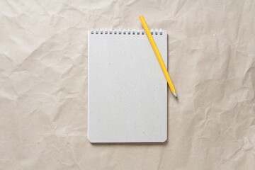 Gray notepad with white coiled spring and pencil on a background of beige crumpled craft paper. With empty space for text and design