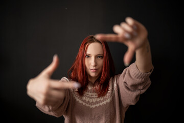 a beautiful and natural smiling red-haired skinny girl in a knitted sweater makes an imaginary lens or camera frame gesture with her fingers and looks through it at the viewer - Powered by Adobe