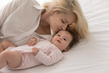 Happy loving mom kissing sweet few month baby with care, affection, tenderness, resting on bed...