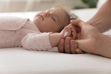 Young parents holding tiny hand of adorable baby girl sleeping on bed. Few month kid resting on mattress with mom and dad touching child arm. Close up of hands, cropped shot. Childcare, family