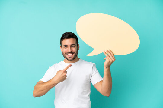 Young handsome caucasian man isolated on blue background holding an empty speech bubble and pointing it