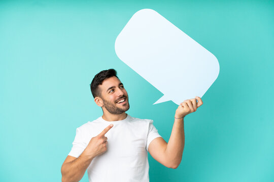 Young handsome caucasian man isolated on blue background holding an empty speech bubble and pointing it