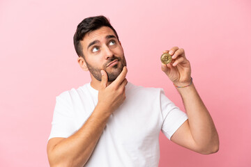 Young man holding a Bitcoin isolated on pink background having doubts