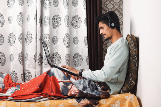 Young bearded Indian boy typing on laptop computer with smartwatch on wrist and earphone or headphone in ear with blue tshirt and red bedsheet isolated on textured curtain background